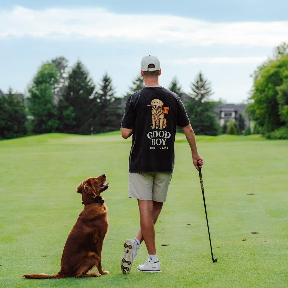 A man on a golf course seen from behind, wearing a black t-shirt with a golden retriever graphic and the text 'GOOD BOY GOLD CLUB', khaki shorts, and a white cap. A brown dog sits on the grass beside him, and he holds a golf club.