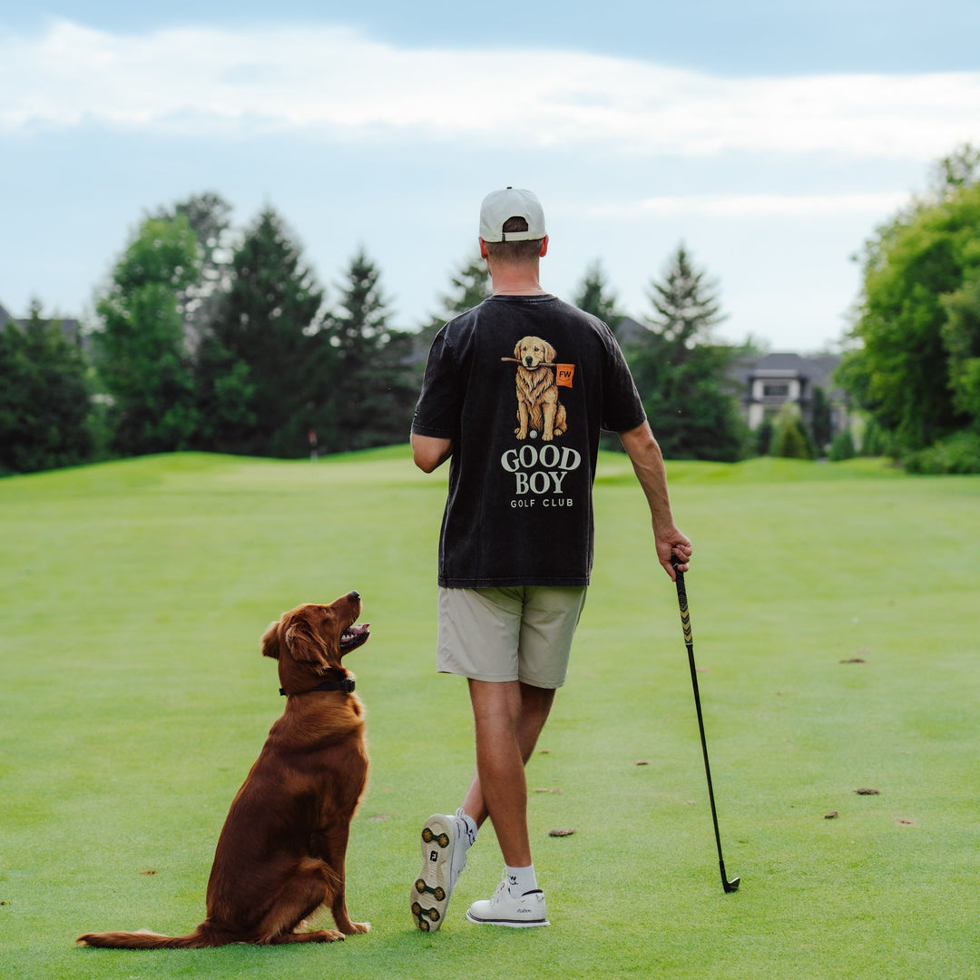 A man on a golf course seen from behind, wearing a black t-shirt with a golden retriever graphic and the text &